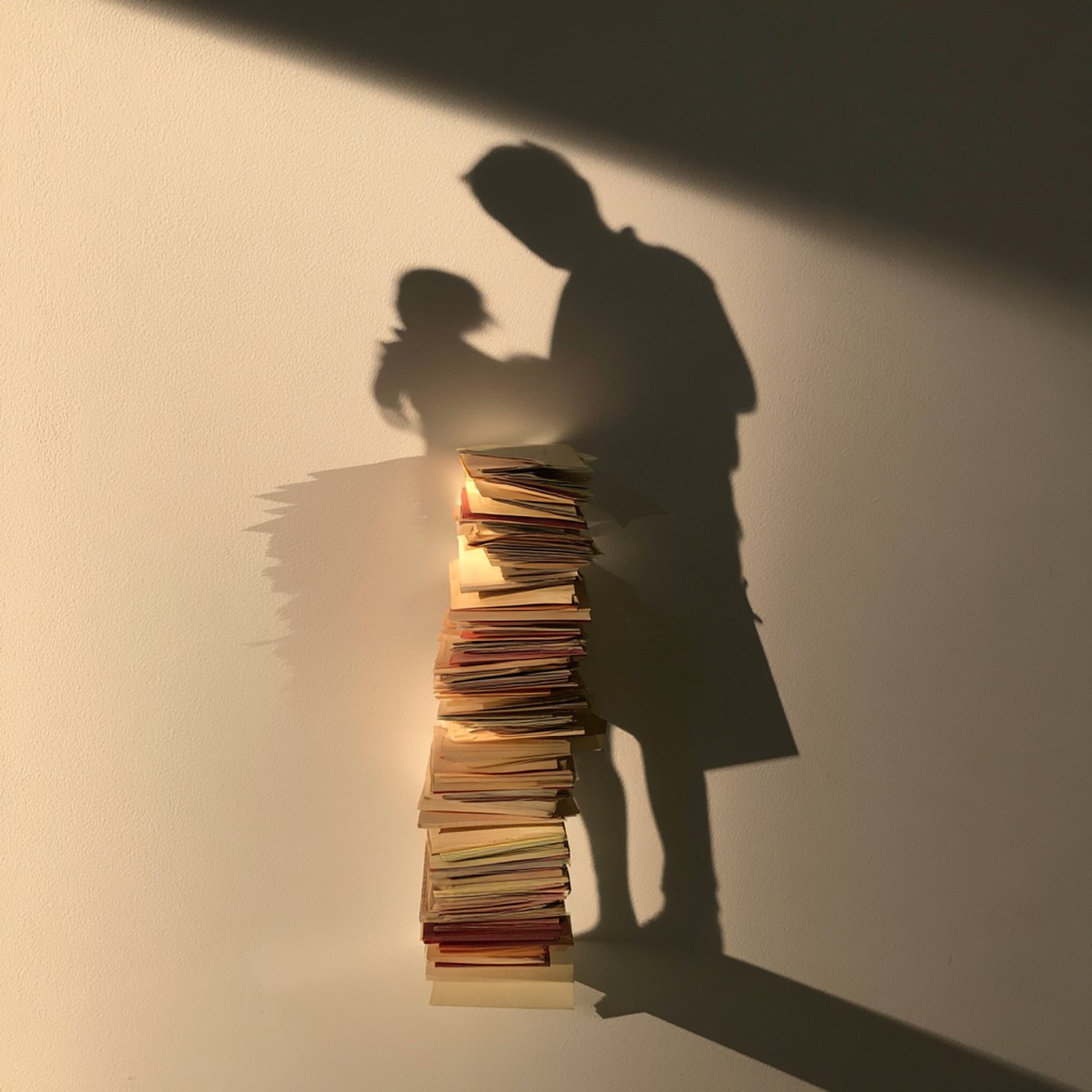 A stack of pulled record-cards stands on a pale surface under a single warm light, throwing a long shadow on the wall that resolves into the silhouette of an adult holding a small child